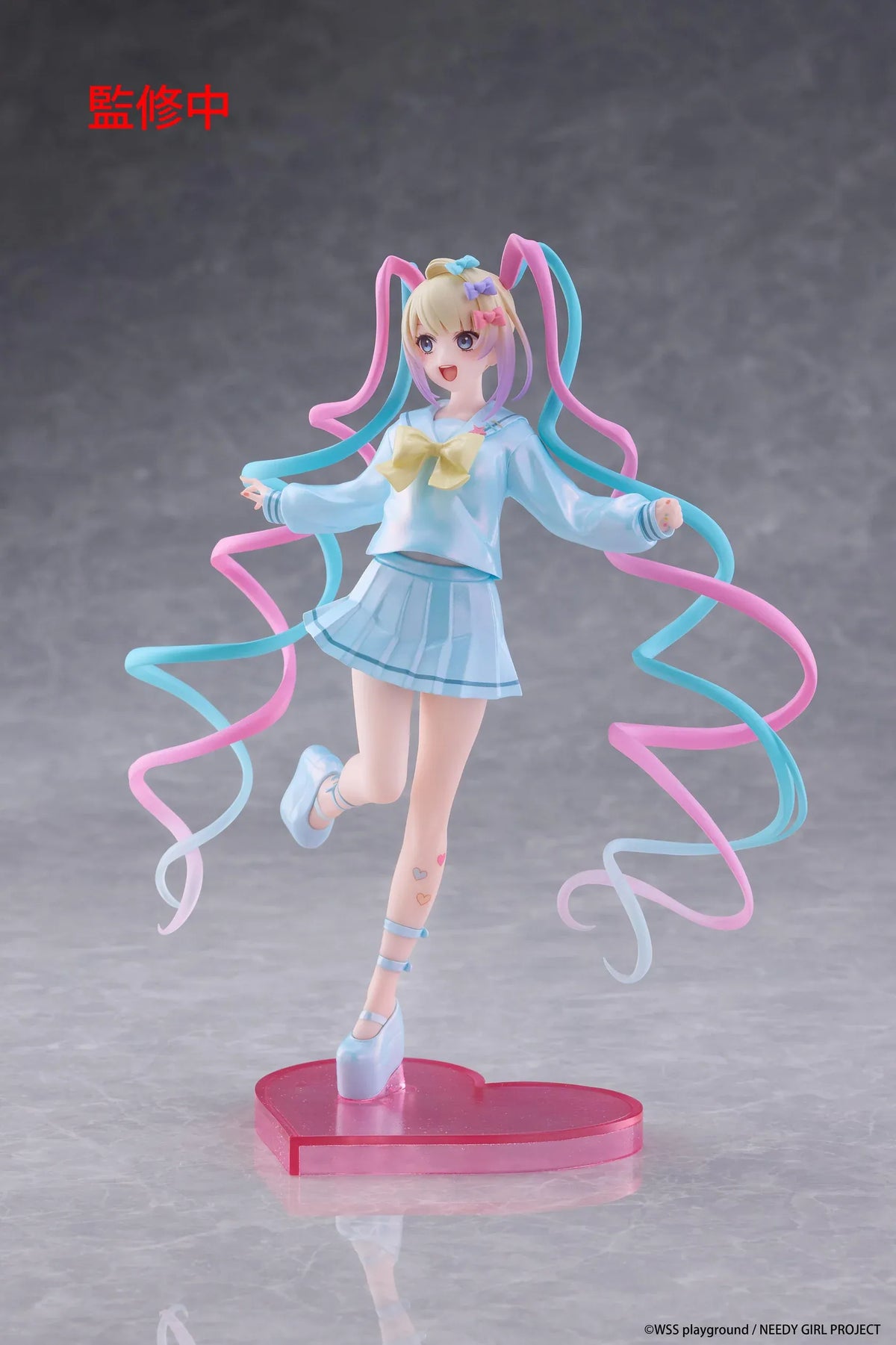 Needy Girl Overdose OMGKawaiiAngel Coreful Figure featuring a blonde-haired anime girl with blue eyes, long pink and blue twin-tails, a pastel-blue sailor-style outfit, yellow bow, and matching shoes posed on a pink heart-shaped base.