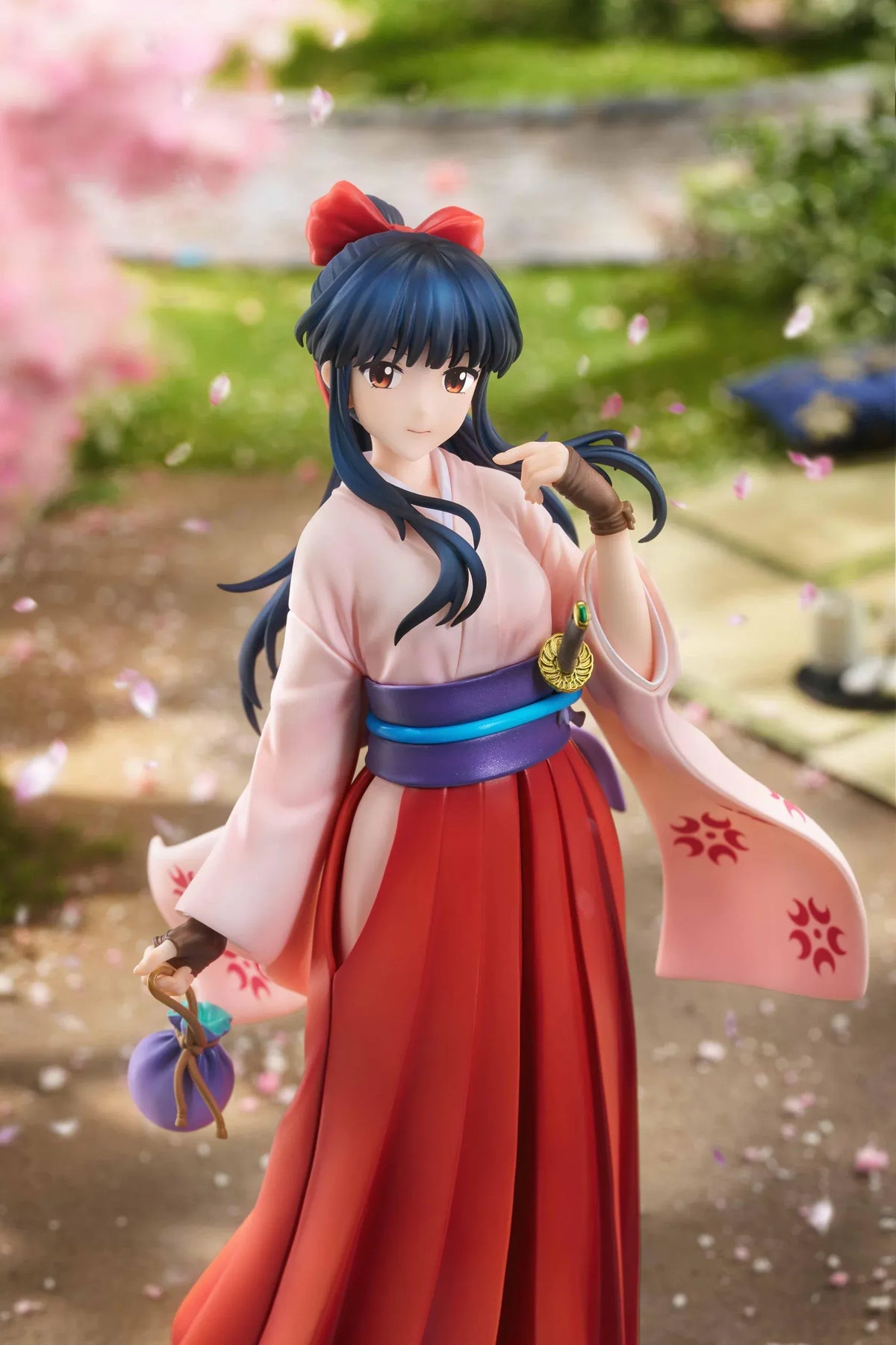 Sakura Wars Sakura Shinguji 1/7 Scale Figure featuring a dark blue-haired anime girl with brown eyes, a red hair ribbon, pink kimono-style top, purple obi, flowing red hakama skirt, sword accessory, and purple pouch on a transparent circular base.