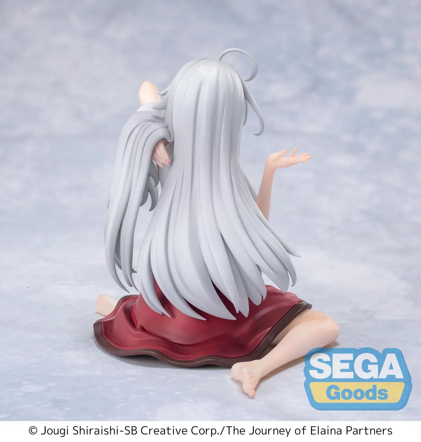 Figure of Elaina from Wandering Witch: The Journey of Elaina sitting on a bed in a white blouse and red skirt, with long silver hair and books beside her, in a sunlit room.