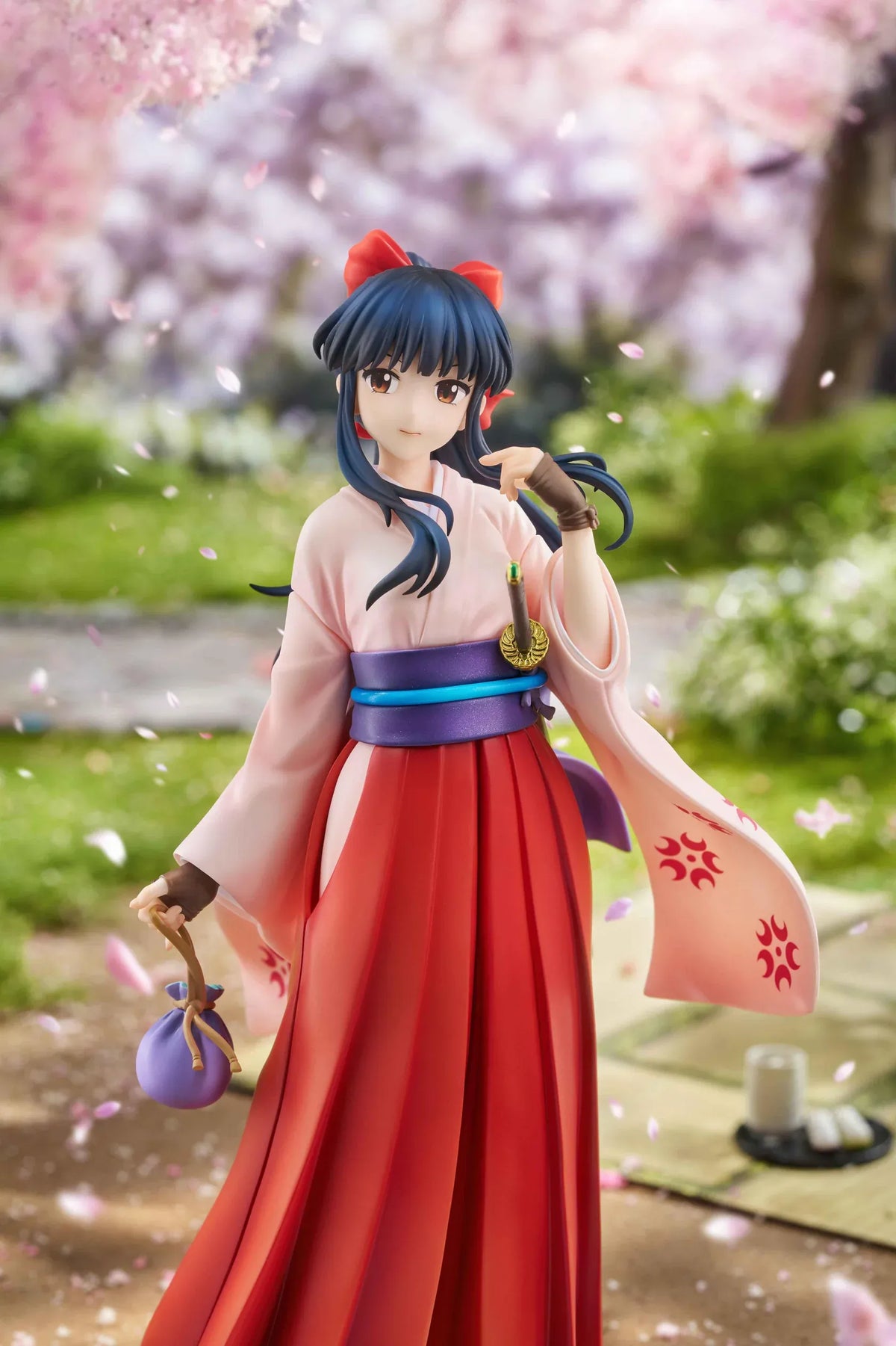 Sakura Wars Sakura Shinguji 1/7 Scale Figure featuring a dark blue-haired anime girl with brown eyes, a red hair ribbon, pink kimono-style top, purple obi, flowing red hakama skirt, sword accessory, and purple pouch on a transparent circular base.