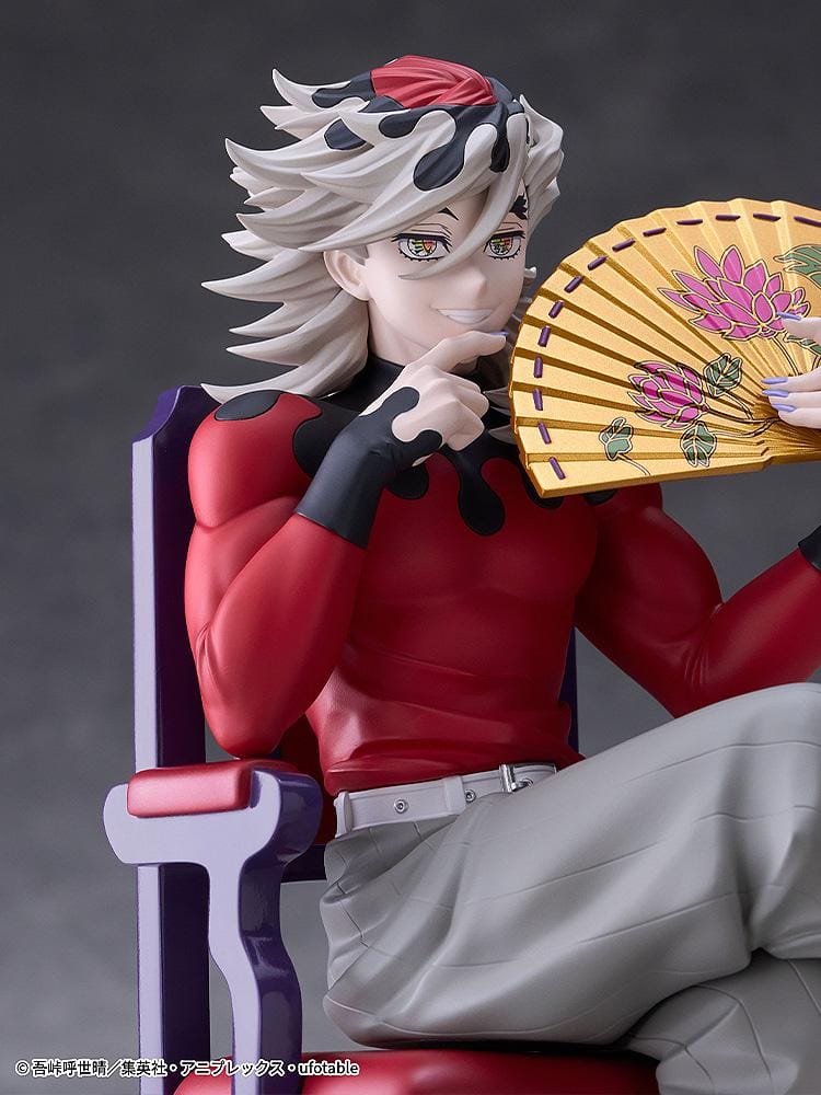 Doma figure seated cross-legged on a purple chair with red cushions, holding a gold floral fan and wearing a red top with gray trousers.