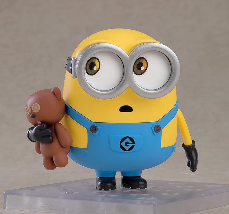 A Nendoroid figure of Bob, the beloved character from the Minions, in his characteristic yellow and blue attire. He's shown with a joyous expression, wearing his typical silver goggles, and accompanied by a teddy bear. In some poses, Bob is waving enthusiastically, while in others, he dons a king's crown and a cape, or holds a bedtime teddy bear, showcasing his playful and lovable personality.