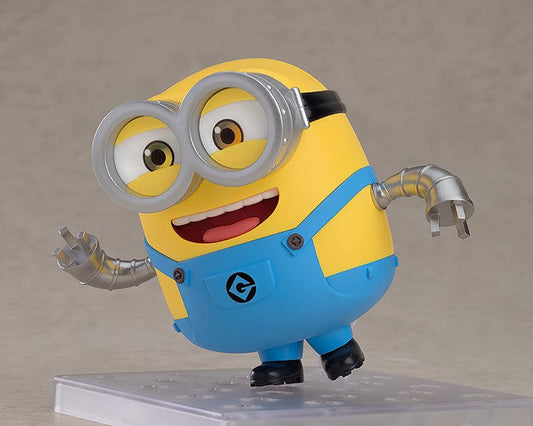 A Nendoroid figure of Bob, the beloved character from the Minions, in his characteristic yellow and blue attire. He's shown with a joyous expression, wearing his typical silver goggles, and accompanied by a teddy bear. In some poses, Bob is waving enthusiastically, while in others, he dons a king's crown and a cape, or holds a bedtime teddy bear, showcasing his playful and lovable personality.