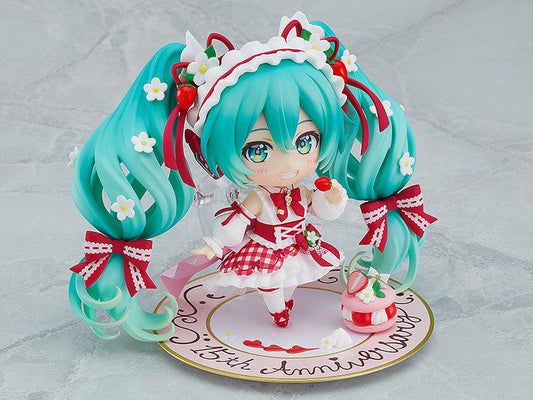 “Nendoroid No.1939 Hatsune Miku (15th Anniversary Ver.) re-run—teal twin-tails with daisies and red bows, frilly dress, holding a strawberry and gold fork, macaron on plate base.”
