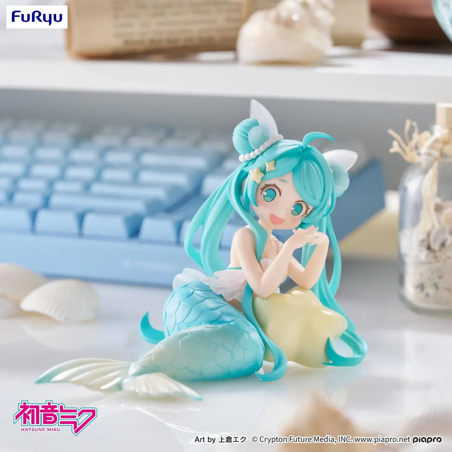 Pastel mermaid Hatsune Miku desktop fairy figure lounging on a star cushion with a mint-green tail and twin-tails, displayed on a desk with seashells and a keyboard in the background.