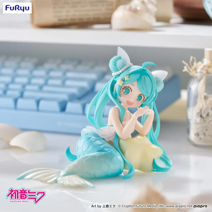 Pastel mermaid Hatsune Miku desktop fairy figure lounging on a star cushion with a mint-green tail and twin-tails, displayed on a desk with seashells and a keyboard in the background.