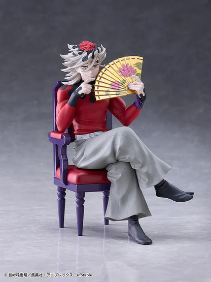 Doma figure seated cross-legged on a purple chair with red cushions, holding a gold floral fan and wearing a red top with gray trousers.