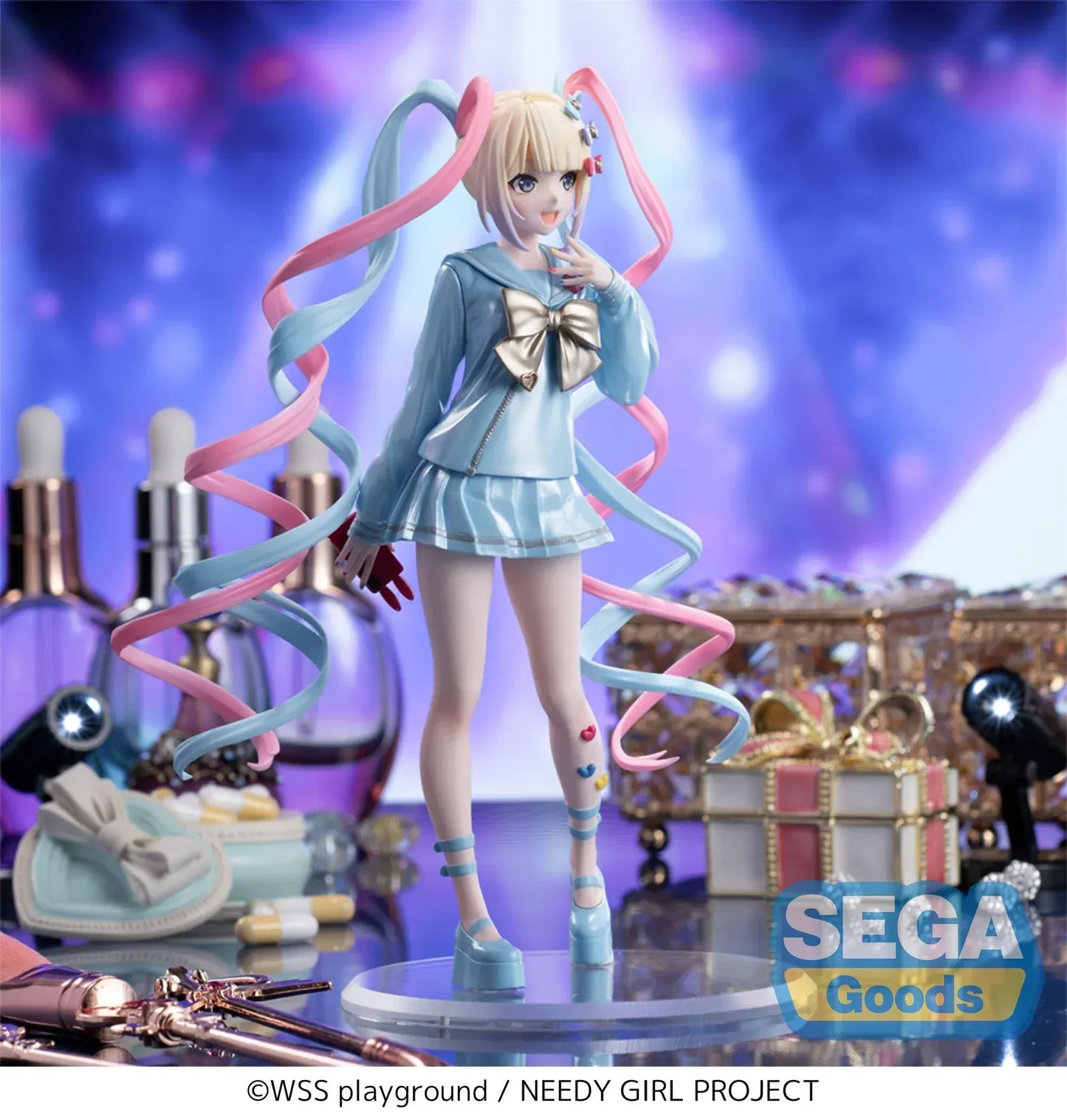 Luminasta OMGKawaiiAngel figure from Needy Streamer Overload standing on a clear base, wearing a pastel blue school outfit with gold bow and long pink-and-blue spiral twin tails, surrounded by makeup and accessories.