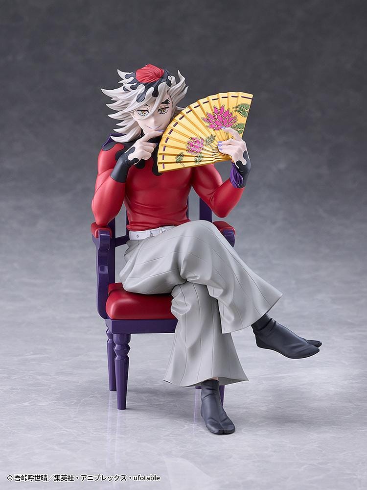 Doma figure seated cross-legged on a purple chair with red cushions, holding a gold floral fan and wearing a red top with gray trousers.