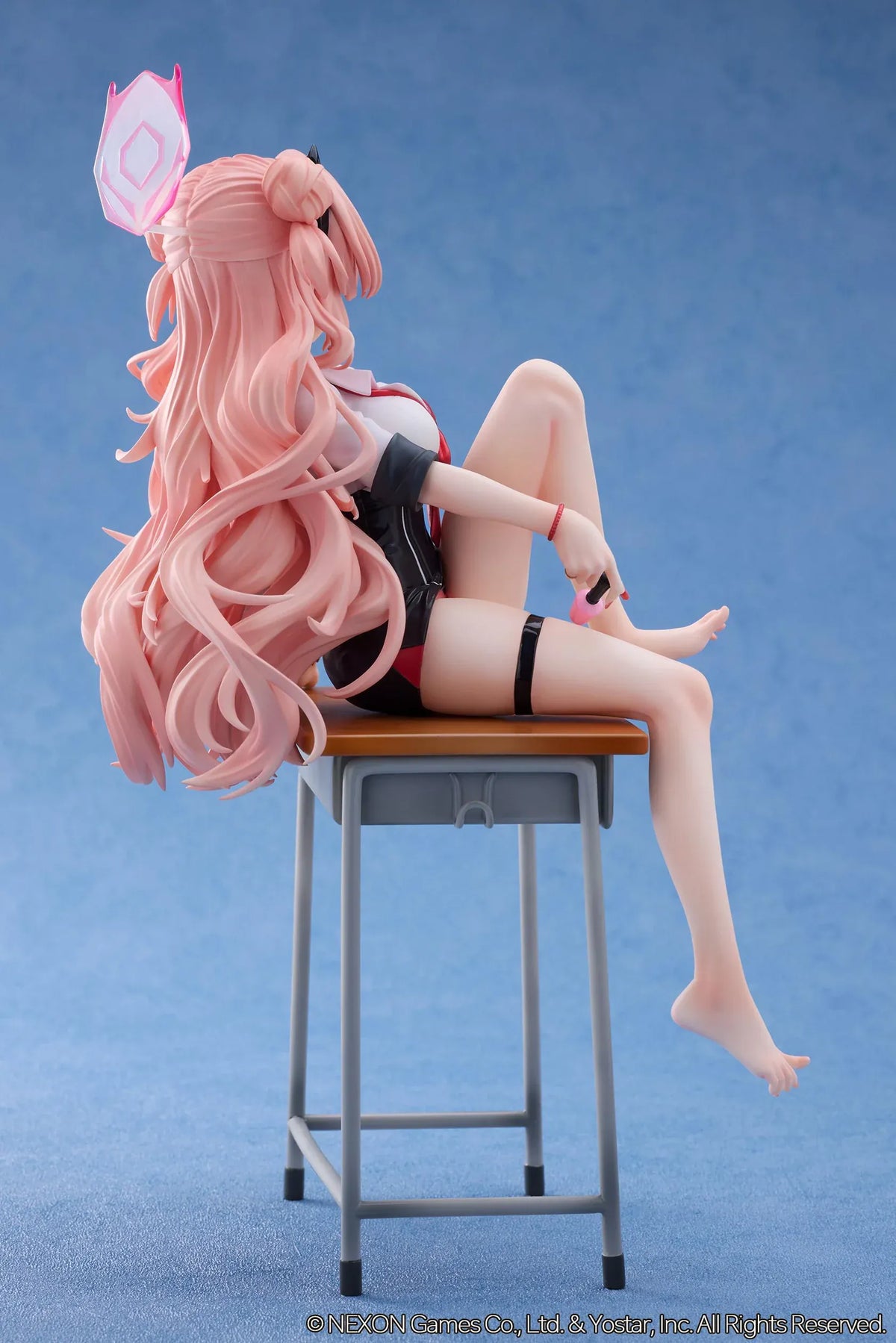 Blue Archive Kirara figure with long pink hair, black horns, and a pink halo, seated on a classroom desk beside nail polish bottles.