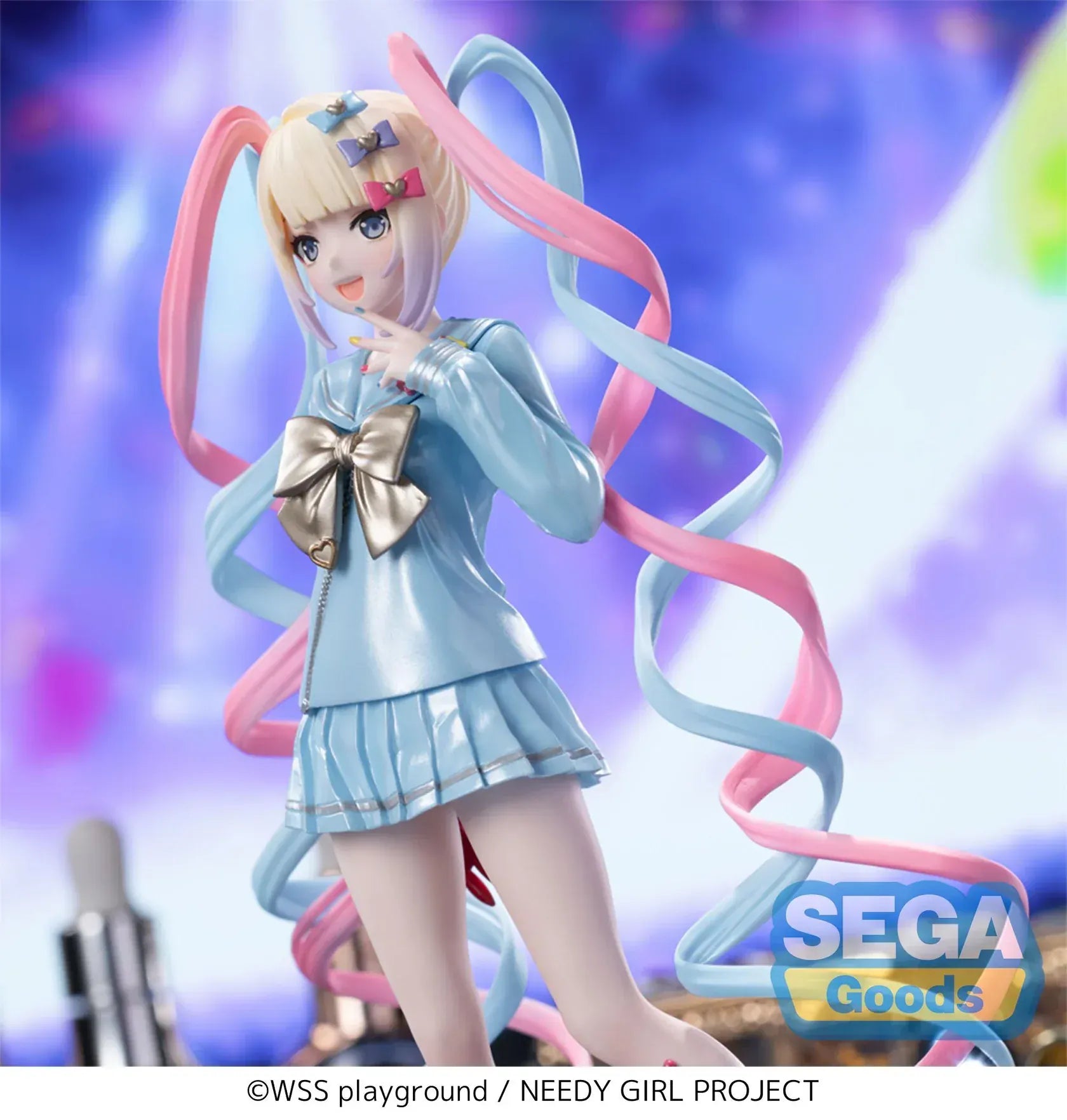 Luminasta OMGKawaiiAngel figure from Needy Streamer Overload standing on a clear base, wearing a pastel blue school outfit with gold bow and long pink-and-blue spiral twin tails, surrounded by makeup and accessories.