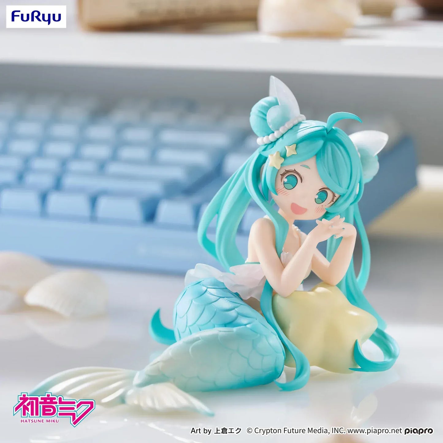 Pastel mermaid Hatsune Miku desktop fairy figure lounging on a star cushion with a mint-green tail and twin-tails, displayed on a desk with seashells and a keyboard in the background.