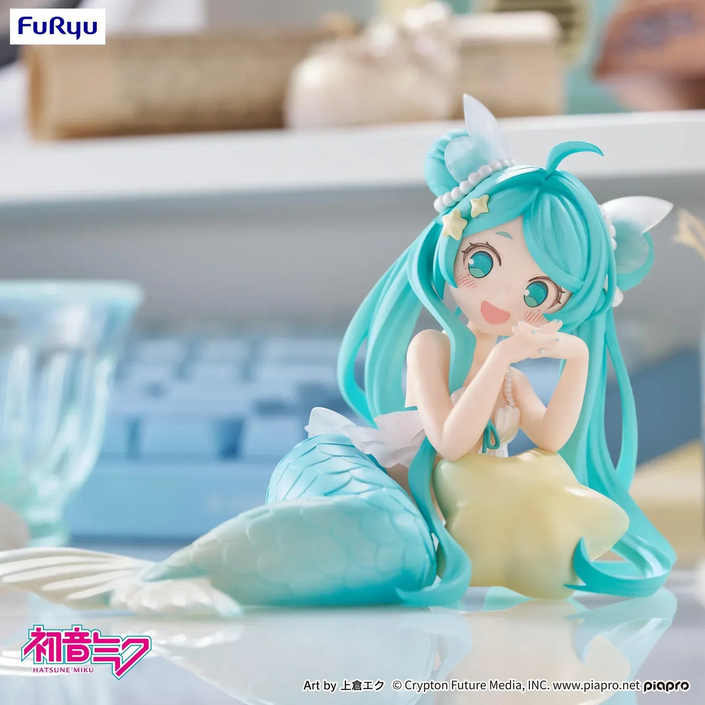 Pastel mermaid Hatsune Miku desktop fairy figure lounging on a star cushion with a mint-green tail and twin-tails, displayed on a desk with seashells and a keyboard in the background.