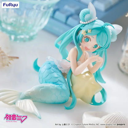 Pastel mermaid Hatsune Miku desktop fairy figure lounging on a star cushion with a mint-green tail and twin-tails, displayed on a desk with seashells and a keyboard in the background.