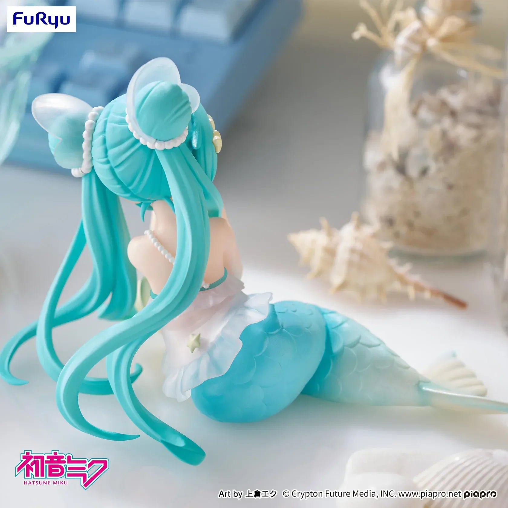 Pastel mermaid Hatsune Miku desktop fairy figure lounging on a star cushion with a mint-green tail and twin-tails, displayed on a desk with seashells and a keyboard in the background.