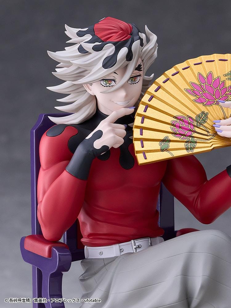 Doma figure seated cross-legged on a purple chair with red cushions, holding a gold floral fan and wearing a red top with gray trousers.