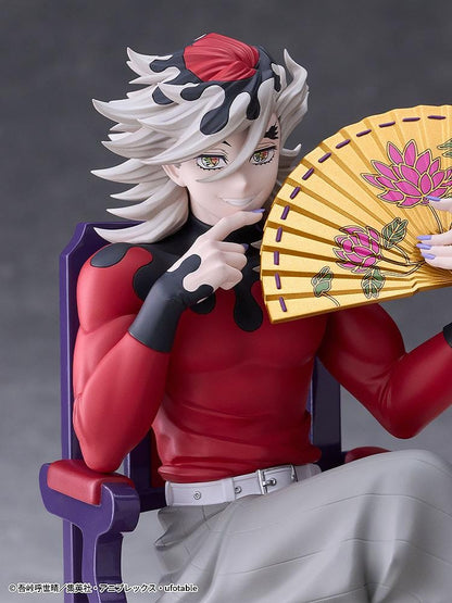 Doma figure seated cross-legged on a purple chair with red cushions, holding a gold floral fan and wearing a red top with gray trousers.