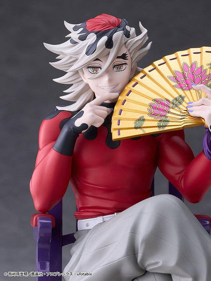 Doma figure seated cross-legged on a purple chair with red cushions, holding a gold floral fan and wearing a red top with gray trousers.