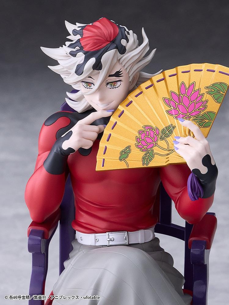 Doma figure seated cross-legged on a purple chair with red cushions, holding a gold floral fan and wearing a red top with gray trousers.