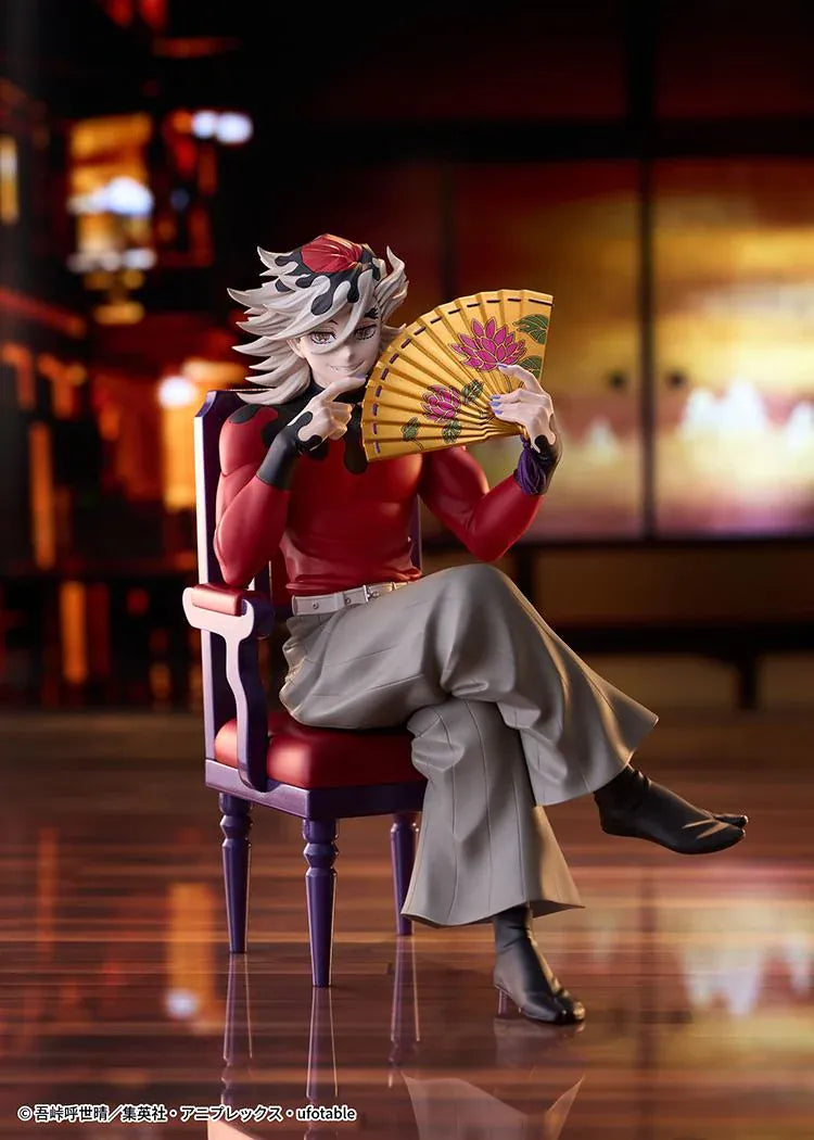 Doma figure seated cross-legged on a purple chair with red cushions, holding a gold floral fan and wearing a red top with gray trousers.