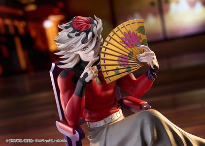 Doma figure seated cross-legged on a purple chair with red cushions, holding a gold floral fan and wearing a red top with gray trousers.