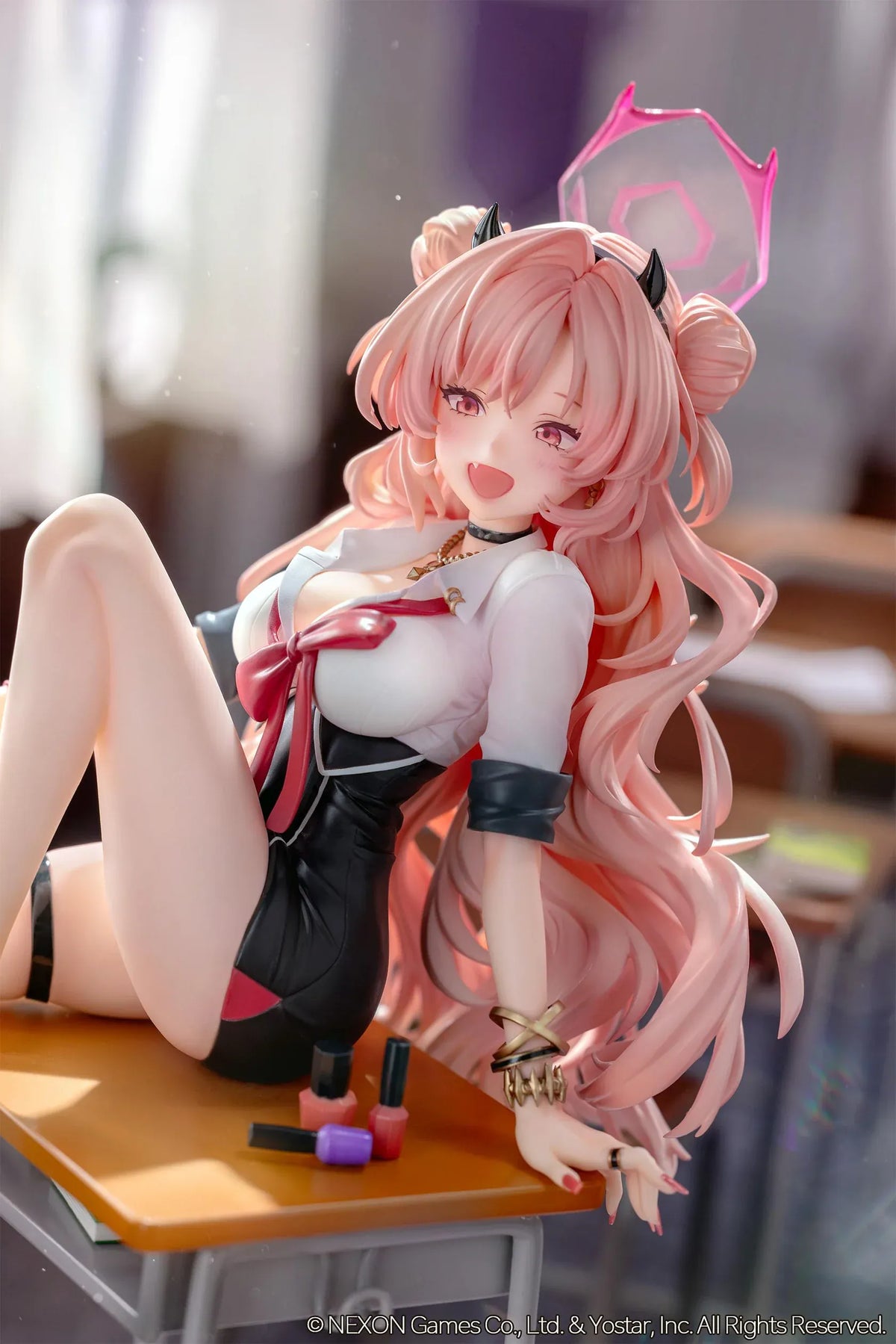 Blue Archive Kirara figure with long pink hair, black horns, and a pink halo, seated on a classroom desk beside nail polish bottles.