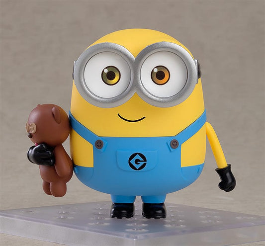 A Nendoroid figure of Bob, the beloved character from the Minions, in his characteristic yellow and blue attire. He's shown with a joyous expression, wearing his typical silver goggles, and accompanied by a teddy bear. In some poses, Bob is waving enthusiastically, while in others, he dons a king's crown and a cape, or holds a bedtime teddy bear, showcasing his playful and lovable personality.