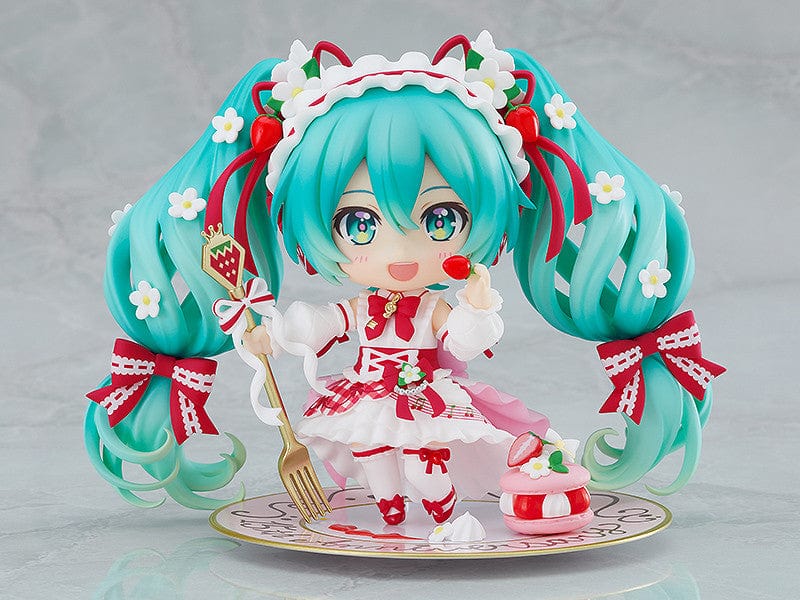 “Nendoroid No.1939 Hatsune Miku (15th Anniversary Ver.) re-run—teal twin-tails with daisies and red bows, frilly dress, holding a strawberry and gold fork, macaron on plate base.”

