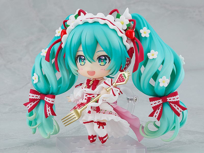 “Nendoroid No.1939 Hatsune Miku (15th Anniversary Ver.) re-run—teal twin-tails with daisies and red bows, frilly dress, holding a strawberry and gold fork, macaron on plate base.”

