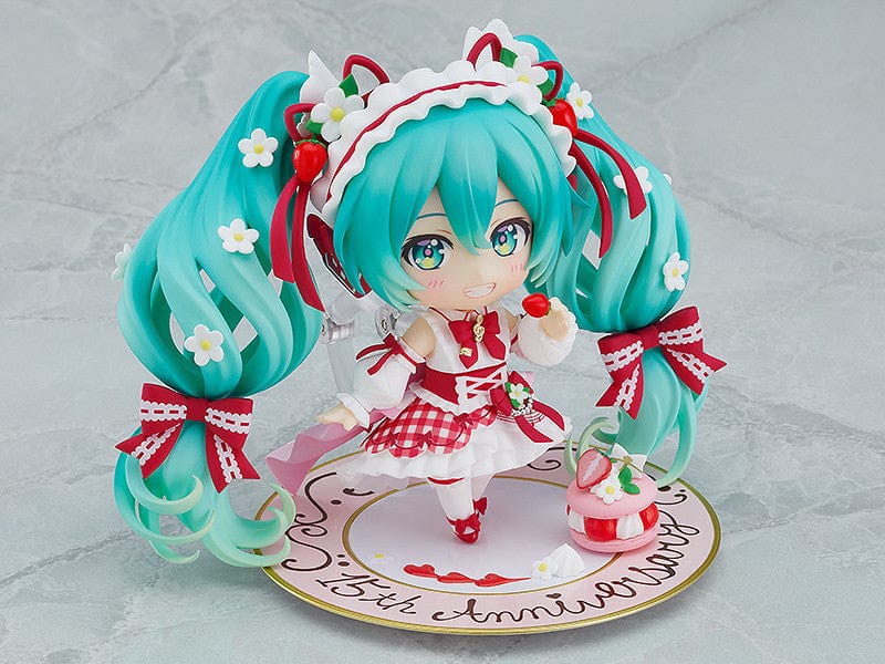 “Nendoroid No.1939 Hatsune Miku (15th Anniversary Ver.) re-run—teal twin-tails with daisies and red bows, frilly dress, holding a strawberry and gold fork, macaron on plate base.”

