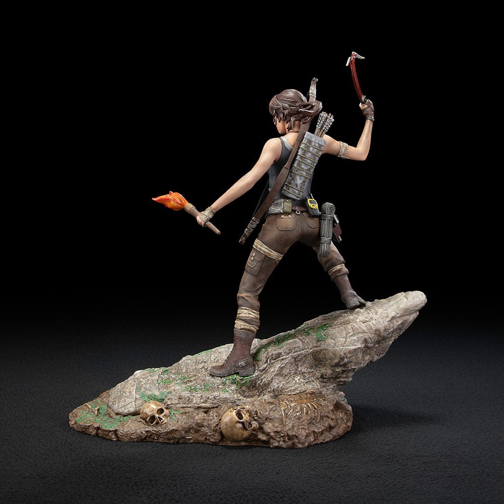 Tomb Raider Lara Croft (Survivor Era) Statue holding climbing axe and torch, in rugged survival gear on rocky base with foliage and skull